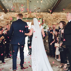 Peut inclure: C&eacute;r&eacute;monie de mariage o&ugrave; les mari&eacute;s marchent dans une all&eacute;e, sous une pluie de p&eacute;tales de roses. La mari&eacute;e porte une robe et un voile en dentelle blanche, le mari&eacute; un costume bleu marine. La c&eacute;r&eacute;monie se d&eacute;roule dans un lieu rustique avec des murs en briques apparentes.