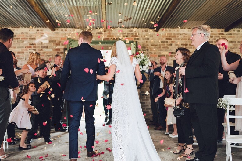 Peut inclure: C&eacute;r&eacute;monie de mariage o&ugrave; les mari&eacute;s marchent dans une all&eacute;e, sous une pluie de p&eacute;tales de roses. La mari&eacute;e porte une robe et un voile en dentelle blanche, le mari&eacute; un costume bleu marine. La c&eacute;r&eacute;monie se d&eacute;roule dans un lieu rustique avec des murs en briques apparentes.