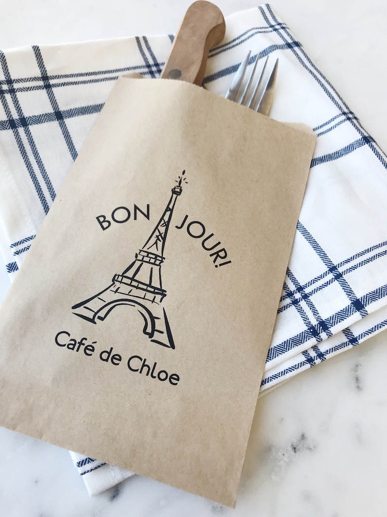 May include: A brown paper bag with a black and white illustration of the Eiffel Tower and the text "Bon Jour! Cafe de Chloe" printed on it. The bag is folded and placed on top of a white and blue checkered cloth napkin.
