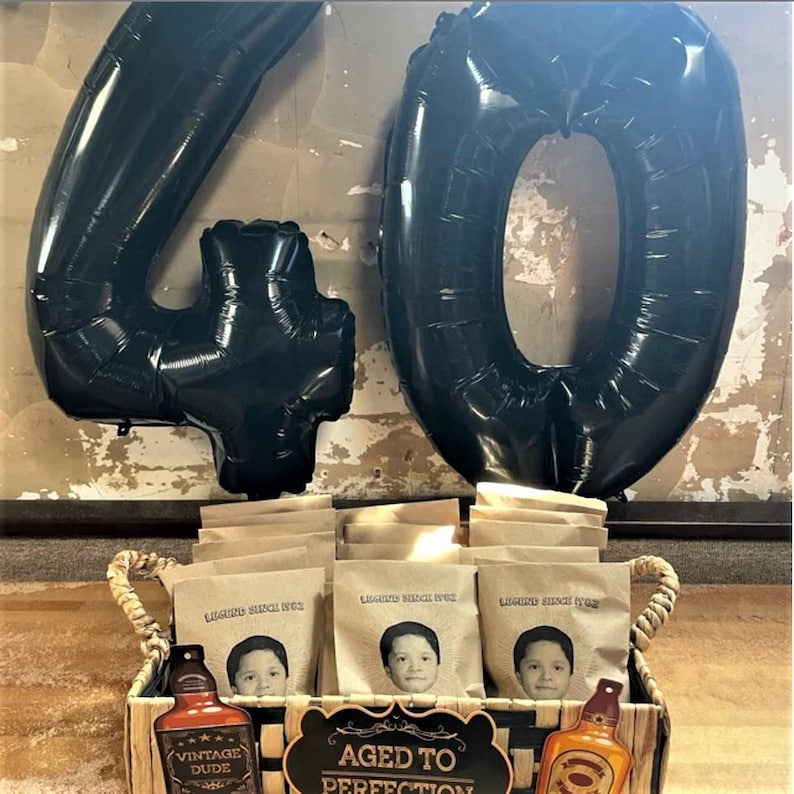 Puede incluir: Globos negros con el n&uacute;mero 40 con una cesta de bolsas de papel marr&oacute;n con una imagen de una persona en cada bolsa. Las bolsas tienen el texto "Liquor Since 1982". La cesta tambi&eacute;n tiene un cartel que dice "Aged to Perfection" y una botella con el texto "Vintage Dude" en ella.