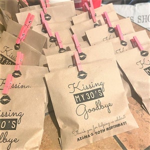 Puede incluir: Bolsas de papel marr&oacute;n con pinzas de ropa rosas y texto negro que dice "Kissing My 30's Goodbye" y "&iexcl;Gracias por ayudarnos a celebrar el 40 cumplea&ntilde;os de Aisha!"