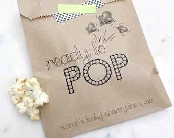 May include: A brown paper bag filled with popcorn, with the words "ready to POP" printed in black. The bag is decorated with a black and white polka dot tape and a small piece of green tape. The words "sarah's baby shower june 3, 2017" are printed at the bottom.