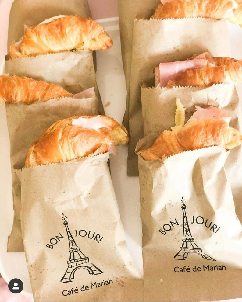 May include: Brown paper bags with croissants inside. The bags have a black and white illustration of the Eiffel Tower with the text "Bon Jour! Cafe de Mariah" printed on them.