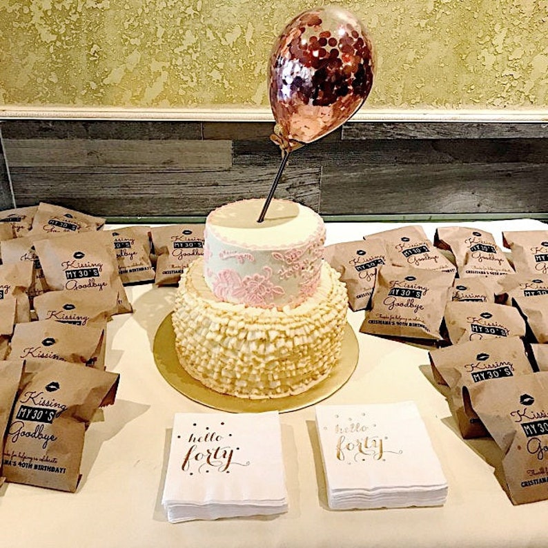 Puede incluir: Un pastel blanco de dos pisos con glaseado rosa y dorado, decorado con un globo de confeti dorado. El pastel est&aacute; rodeado de peque&ntilde;as bolsas de papel marr&oacute;n con el texto "Kissing My 30's Goodbye" impreso en ellas. Tambi&eacute;n hay dos servilletas blancas con el texto "Hello Forty" impreso en ellas.