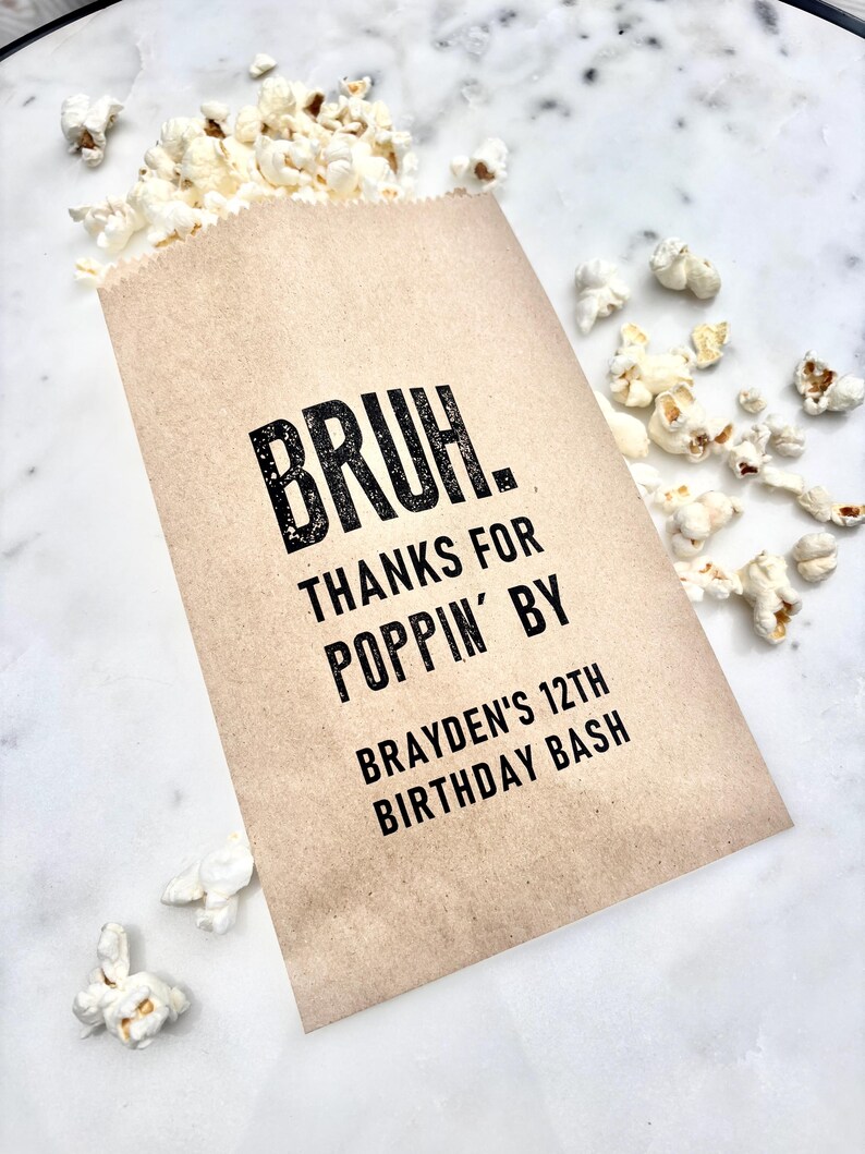 May include: A brown paper popcorn bag with the text "BRUH. THANKS FOR POPPIN' BY BRAYDEN'S 12TH BIRTHDAY BASH" in black. The bag is filled with white popcorn, with some kernels scattered around on a white marble surface.