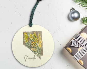 Peut inclure: Une décoration ronde de couleur crème avec une ficelle verte pour l'accrocher. La décoration présente une illustration aquarelle de l'État du Nevada avec des fleurs jaunes. Le texte "Nevada" est imprimé sous l'illustration.