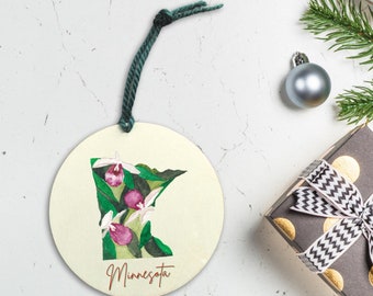 Peut inclure: Une décoration ronde de couleur crème avec une ficelle verte pour l'accrocher. La décoration présente une illustration aquarelle de l'état du Minnesota avec des fleurs de sabot de Vénus roses et des feuilles vertes. Le mot "Minnesota" est écrit en lettres dorées au bas de la décoration.