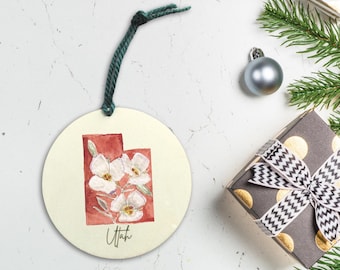 Peut inclure: Ornement de Noël rond avec le contour de l'Utah en rouge et une illustration de fleurs blanches. Le mot "Utah" est écrit en cursive sur un fond crème. L'ornement est suspendu par un cordon vert.