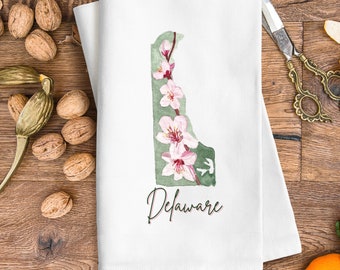 Peut inclure: Une serviette de cuisine blanche avec une illustration aquarelle de l'état du Delaware délimité en vert avec des fleurs roses. Le texte "Delaware" est écrit en cursive sous l'état.