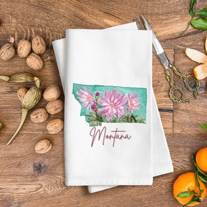 May include: White tea towel with a watercolor illustration of the state of Montana with pink flowers and the word "Montana" in cursive.