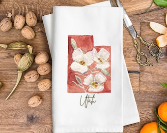 Peut inclure: Serviette de cuisine blanche avec une illustration aquarelle de l'état de l'Utah avec des fleurs blanches et le mot "Utah" en écriture verte.