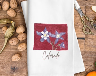 Peut inclure: Une serviette de cuisine blanche avec un fond rouge représentant une illustration aquarelle de fleurs de colombine bleues et blanches. Le texte "Colorado" est écrit en cursive noire sous les fleurs.