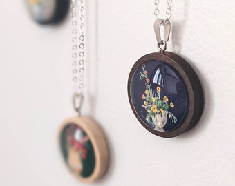 Peut inclure: Un collier en chaîne argentée avec un pendentif rond en bois représentant un motif floral sur fond bleu foncé. Le pendentif est encadré d'une bordure en bois brun foncé.