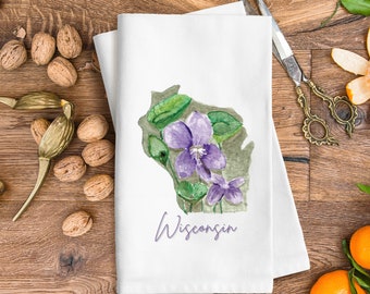 Peut inclure: Une serviette de cuisine blanche avec une illustration aquarelle d'une violette violette en forme de l'état du Wisconsin. Le texte "Wisconsin" est écrit sous la fleur.
