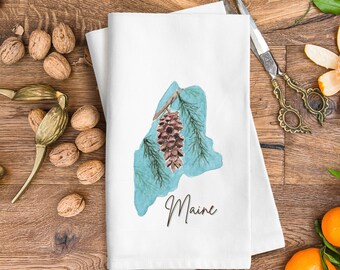 Peut inclure: Une serviette de cuisine blanche avec un contour bleu aquarelle de l'état du Maine. Un cône de pin brun est représenté à l'intérieur du contour. Le texte "Maine" est écrit en cursive brune sous le contour.