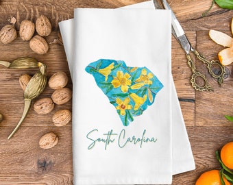 Peut inclure: Une serviette de cuisine blanche avec une illustration aquarelle bleue et jaune de l'état de Caroline du Sud. L'état est délimité en bleu et rempli de fleurs jaunes. Le texte "South Carolina" est écrit en vert sous l'état.