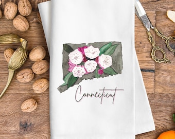 Peut inclure: Une serviette de cuisine blanche avec une illustration aquarelle de l'état du Connecticut avec des fleurs roses et blanches. Le texte "Connecticut" est écrit en cursive sous l'état.