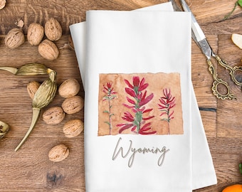Peut inclure: Une serviette de cuisine blanche avec une illustration aquarelle de fleurs rouges d'Indian paintbrush sur un fond brun. Le texte "Wyoming" est imprimé sous l'illustration.