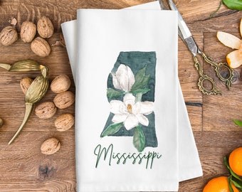 Peut inclure: Une serviette de cuisine blanche avec une illustration aquarelle de l'état du Mississippi avec une fleur de magnolia en forme d'état. Le texte "Mississippi" est écrit en cursive verte sous l'état.