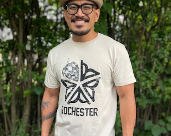 Peut inclure: Une personne portant un t-shirt beige avec un motif noir représentant une fleur de houblon et le mot "ROCHESTER" imprimé dessus. La personne porte un chapeau de paille et des lunettes et sourit.