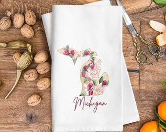 Peut inclure: Une serviette de cuisine blanche avec un motif floral en forme de l'état du Michigan. Le texte "Michigan" est imprimé en bordeaux sous le contour de l'état.