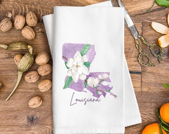 Peut inclure: Une serviette de cuisine blanche avec un contour aquarelle violet de l'état de la Louisiane avec des fleurs de magnolia blanches et des feuilles vertes. Le texte "Louisiana" est écrit en cursive sous le contour de l'état.