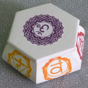 May include: White hexagonal box with seven colorful chakra symbols, including the root chakra, sacral chakra, solar plexus chakra, heart chakra, throat chakra, third eye chakra, and crown chakra. Each chakra symbol is a mandala with a Sanskrit symbol in the center.