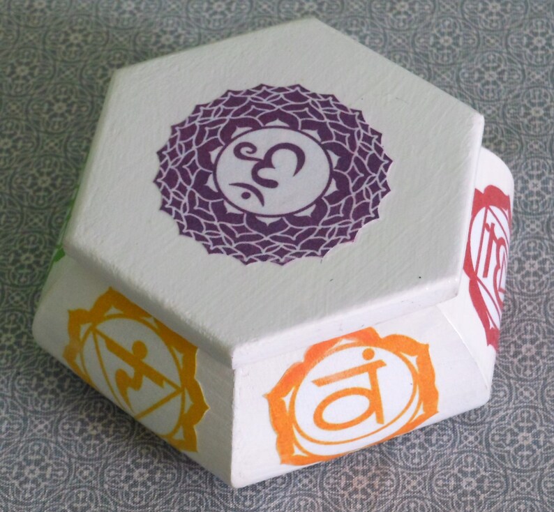 May include: White hexagonal box with seven colorful chakra symbols, including the root chakra, sacral chakra, solar plexus chakra, heart chakra, throat chakra, third eye chakra, and crown chakra. Each chakra symbol is a mandala with a Sanskrit symbol in the center.