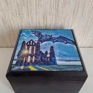 May include: A dark wooden box featuring a detailed illustration on the lid. The artwork shows a gothic cathedral with a bat flying above it against a blue sky. The box has a dark finish.