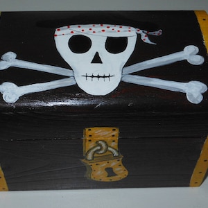 May include: A black wooden treasure chest with a pirate theme. The chest features a white skull with a red-spotted bandana and crossed bones. The edges are trimmed in yellow, and a gold-colored lock is on the front.