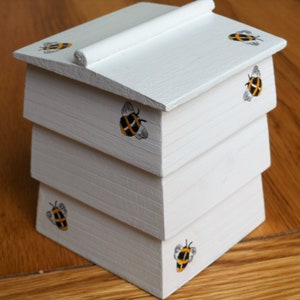 May include: A white wooden beehive with a layered design. The beehive is decorated with black and yellow painted bees.