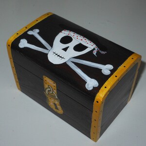 May include: A small, rectangular wooden treasure chest painted dark brown with a gold trim. The lid features a white skull and crossbones design with a red and white polka dot bandana. The chest has a gold clasp.