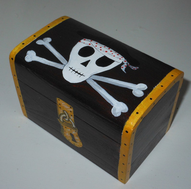 May include: A small, rectangular wooden treasure chest painted dark brown with a gold trim. The lid features a white skull and crossbones design with a red and white polka dot bandana. The chest has a gold clasp.