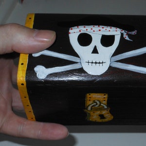 May include: A small, rectangular wooden treasure chest painted black with a skull and crossbones design on the front. The skull has a red and white polka dot bandana. The edges are trimmed with yellow.