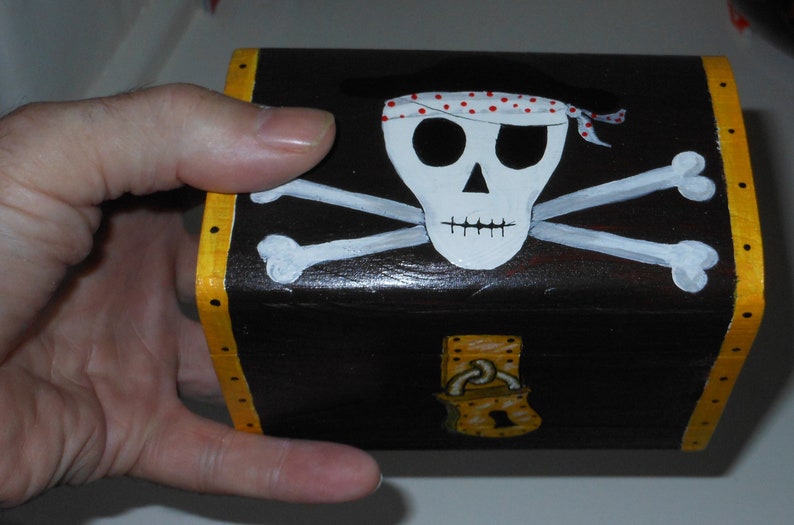 May include: A small, rectangular wooden treasure chest painted black with a skull and crossbones design on the front. The skull has a red and white polka dot bandana. The edges are trimmed with yellow.
