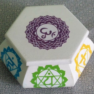 May include: Hexagonal wooden box with a white lid and colorful chakra symbols on each side. The symbols are purple, blue, green, yellow, and orange.