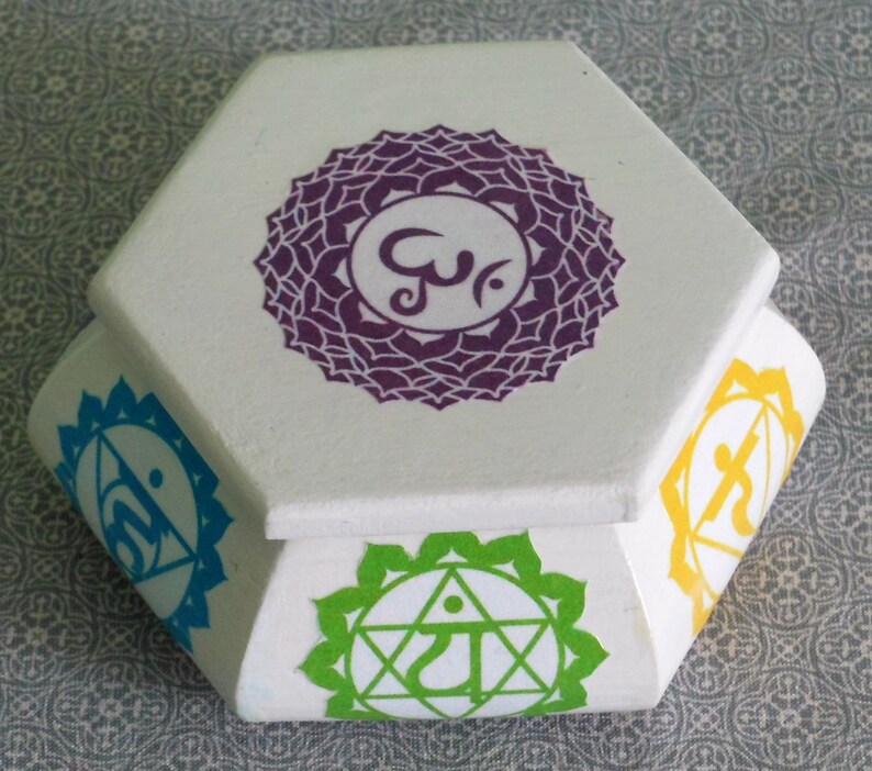 May include: Hexagonal wooden box with a white lid and colorful chakra symbols on each side. The symbols are purple, blue, green, yellow, and orange.
