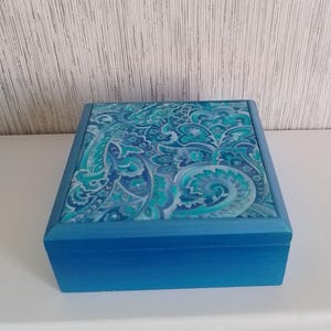 May include: A square, blue wooden box with a patterned lid. The lid features a paisley design in shades of blue and teal. The box itself is a solid, medium blue colour, creating a cohesive and decorative storage item.