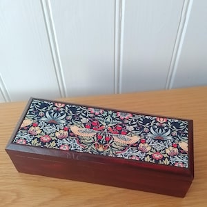 May include: A rectangular wooden box with a dark stain. The lid features a fabric with a floral and bird design in red, yellow, and blue. The box rests on a light wood surface.