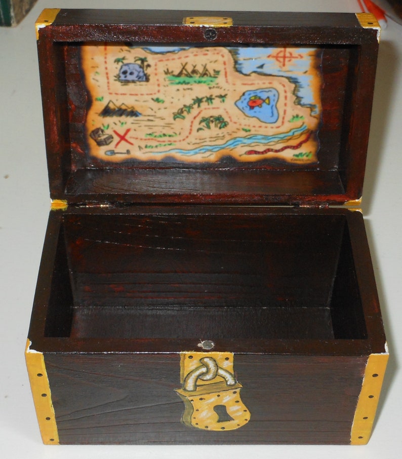 May include: A wooden treasure chest with a dark brown finish, gold accents, and a painted treasure map on the inside lid. The chest is open, revealing the interior. The map features a skull, palm trees, and a red 'X' marking the spot.