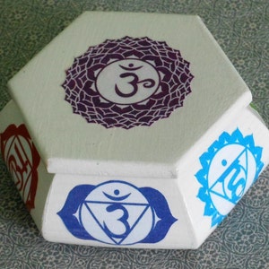 May include: White hexagonal box with a purple, blue, and red chakra symbol on each side. The top of the box has a purple chakra symbol with the Sanskrit word "Om" in the center.
