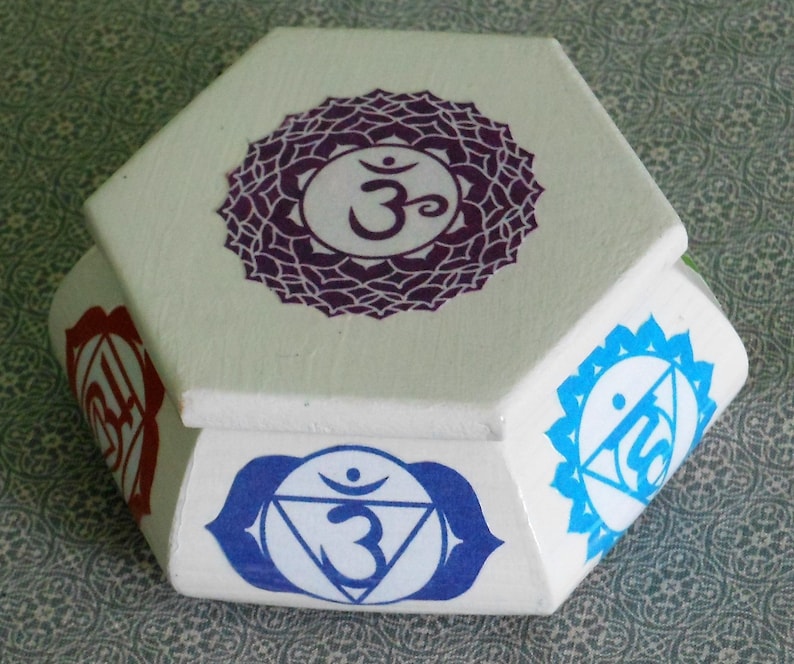 May include: White hexagonal box with a purple, blue, and red chakra symbol on each side. The top of the box has a purple chakra symbol with the Sanskrit word "Om" in the center.