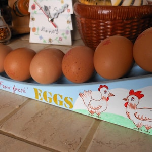 May include: A blue and white egg holder with the words "Farm fresh" and "EGGS" painted on it. Six brown eggs are in the holder. Two painted chickens are on the holder.