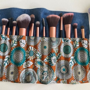 May include: A blue denim makeup brush roll with a floral pattern in shades of orange, teal, and white. The roll is filled with 13 makeup brushes with rose gold handles.