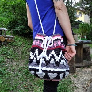 May include: A crocheted drawstring bag with a geometric pattern in pink, white, black, and gray. The bag has a white strap and is worn over the shoulder.