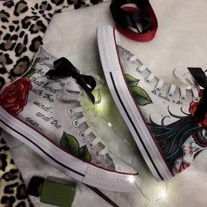 Puede incluir: Un par de zapatos de lona blancos de caña alta con cordones negros y una cinta negra. Los zapatos están pintados a mano con un diseño floral, incluyendo rosas rojas y hojas verdes. Un zapato tiene una cita que dice "Juro que he sido ese pájaro con el viento y la lluvia..." El otro zapato tiene una ilustración en blanco y negro de una mujer con una cara de calavera de azúcar y una rosa roja.