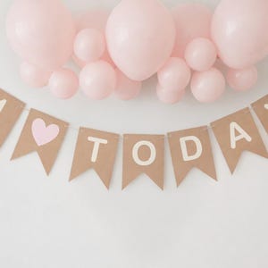 Op de afbeelding: Een bruine papieren banner met de woorden "1 TODAY" in witte letters en een roze hart, onder een groep roze ballonnen. Geschikt voor een eerste verjaardagsfeest.