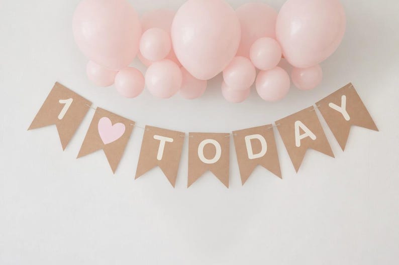Op de afbeelding: Een bruine papieren banner met de woorden "1 TODAY" in witte letters en een roze hart, onder een groep roze ballonnen. Geschikt voor een eerste verjaardagsfeest.