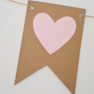 Puede incluir: Una pancarta de papel marr&oacute;n con un coraz&oacute;n rosa recortado. La pancarta est&aacute; enhebrada con un cordel y tiene agujeros perforados en las esquinas superiores.