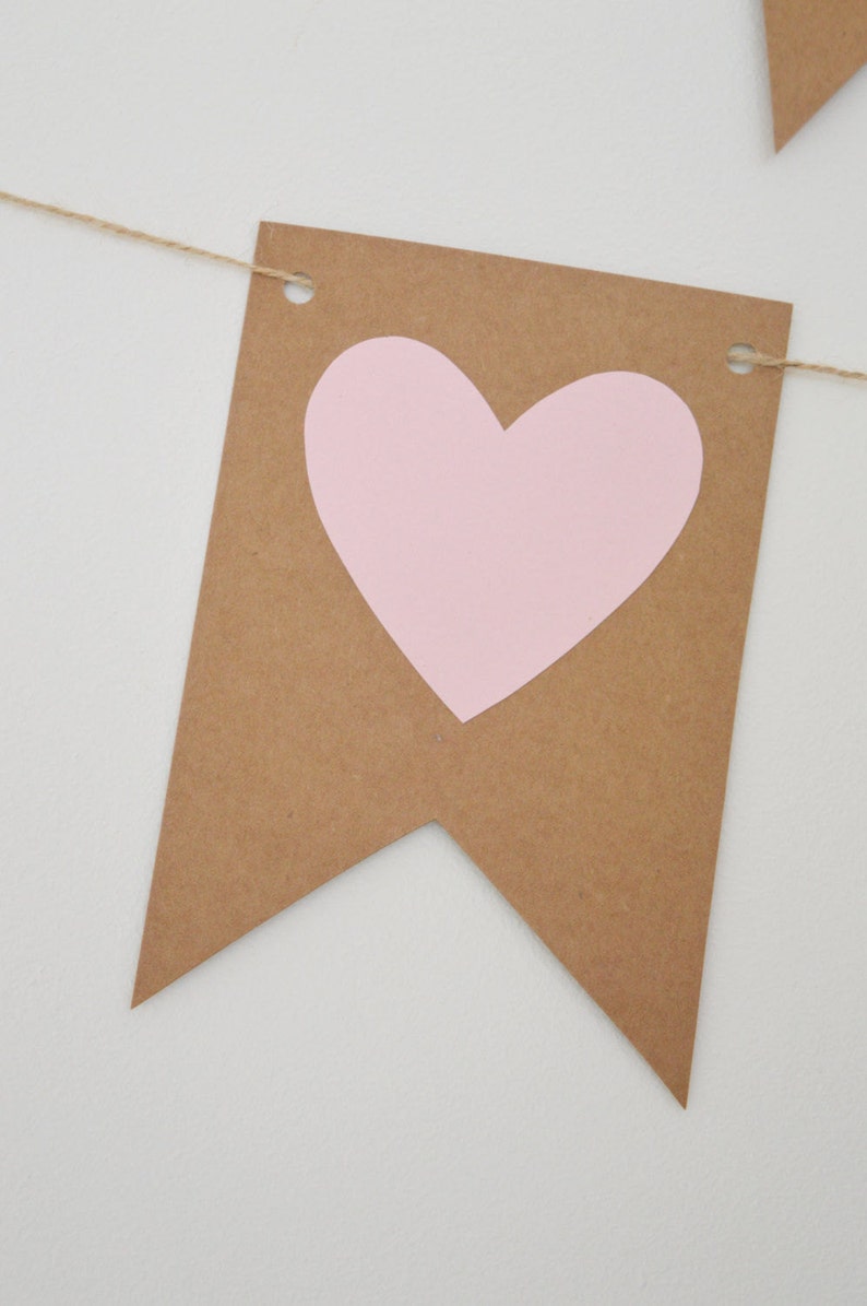 Puede incluir: Una pancarta de papel marr&oacute;n con un coraz&oacute;n rosa recortado. La pancarta est&aacute; enhebrada con un cordel y tiene agujeros perforados en las esquinas superiores.